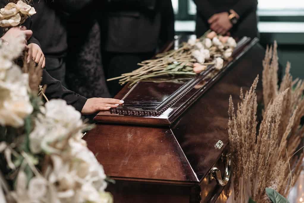 A hand is placed on a dark timber coffin. The coffin is framed by nude and blush floral arrangements. Several long stem roses are placed on top of the coffin. The eulogy is an important part of a funeral, which is depicted in this image.